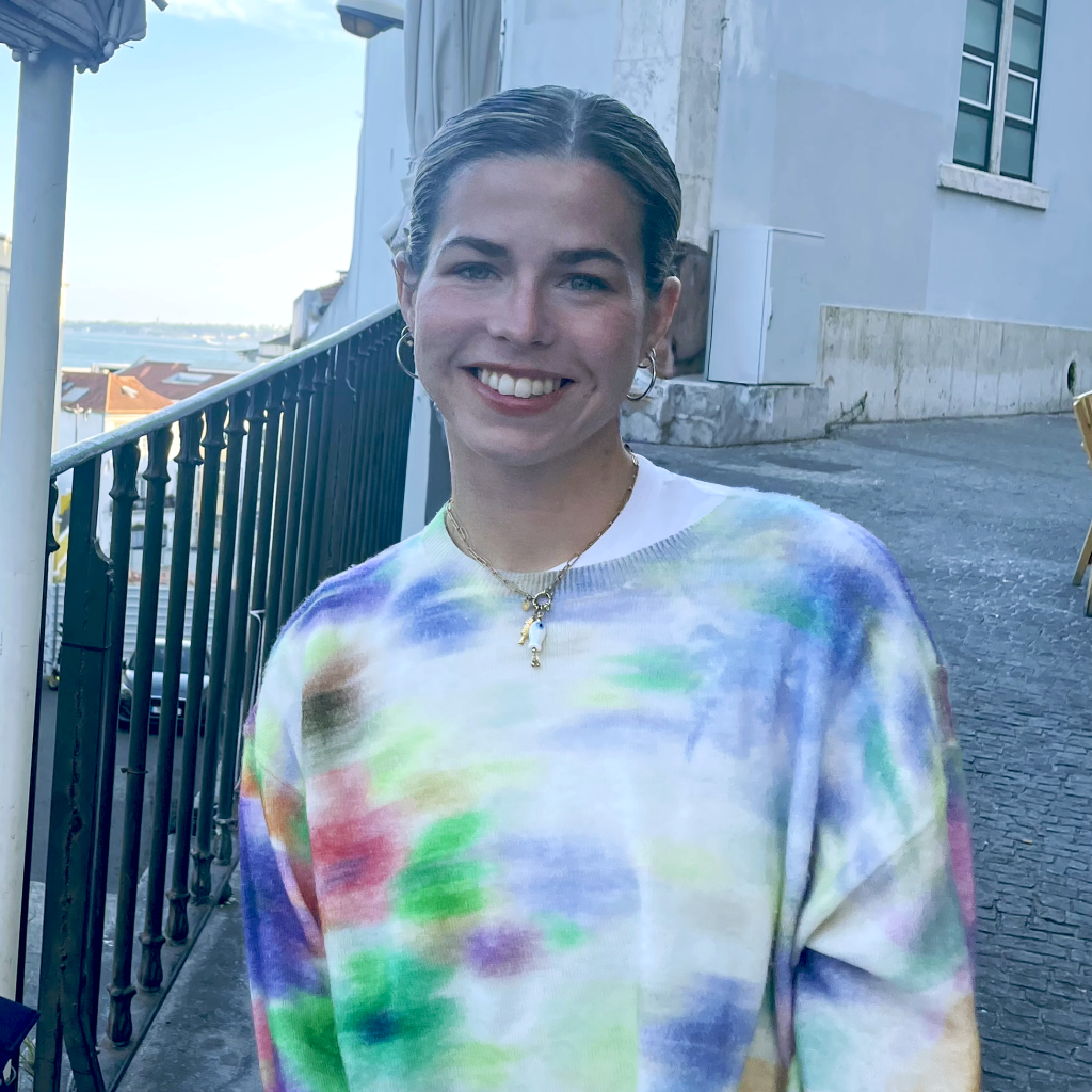A photo of Angela smiling with hair pulled back, wearing small hoop earrings and a pastel tie-dye sweater, standing outdoors beside a railing and light-colored building.